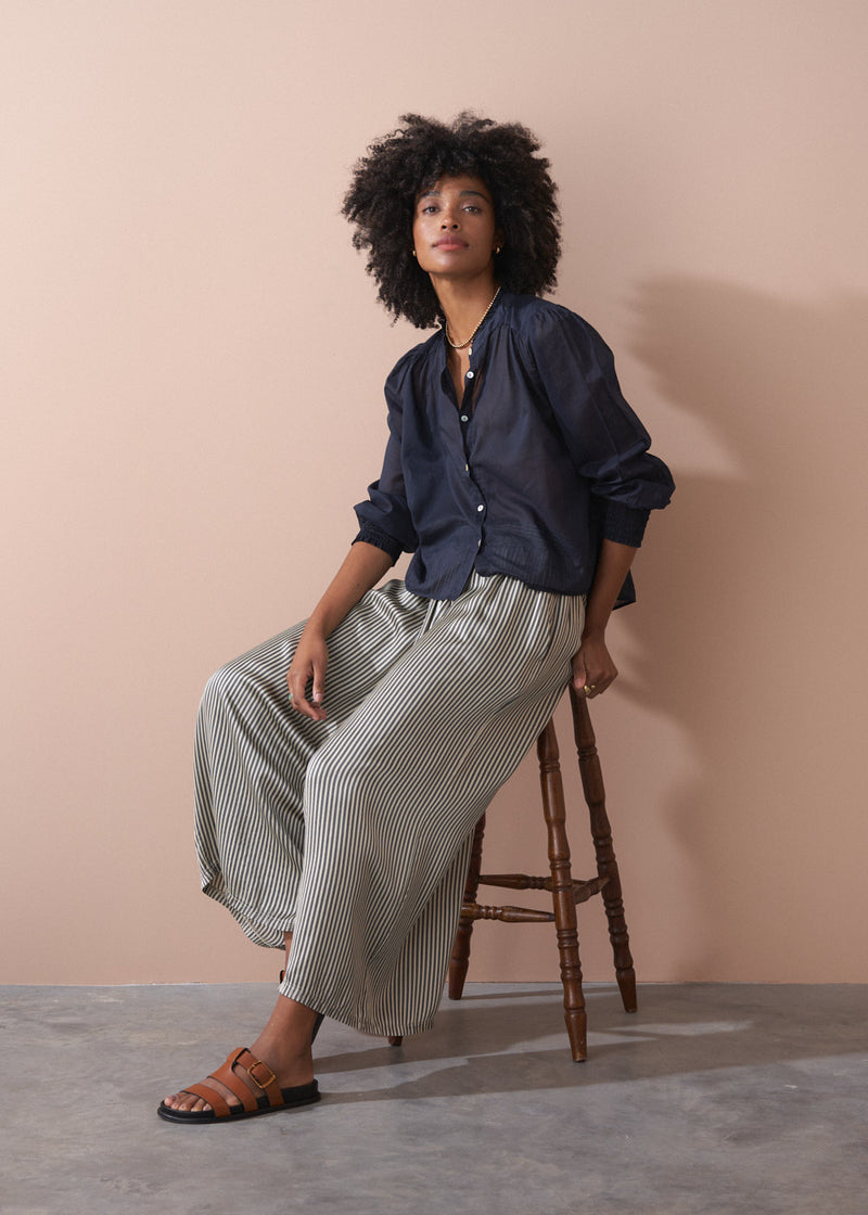 Woman sitting on a stool wearing a navy shirt and striped trousers against a beige background