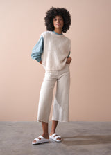 Woman wearing a beige knitted vest and white trosuers against a beige background