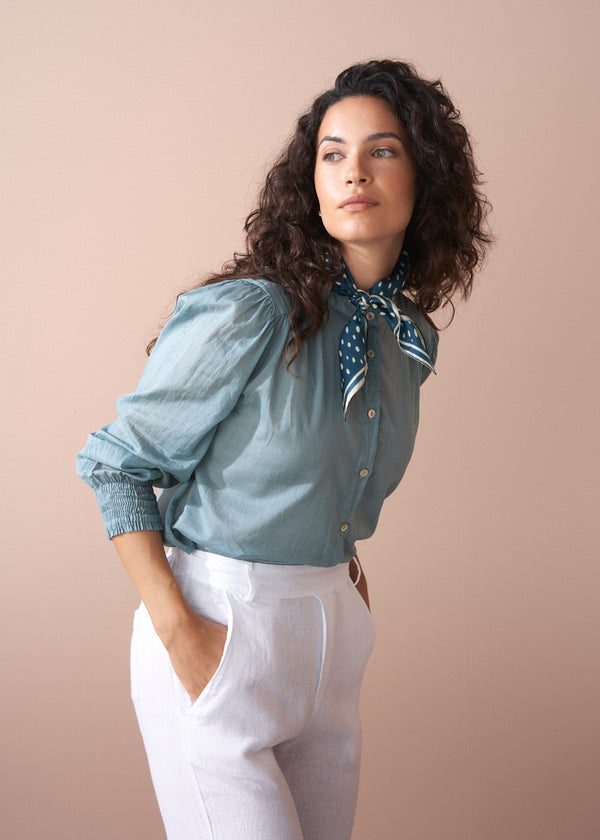 Woman wearing a light blue blouse with puffed sleeves and white pants against a beige background