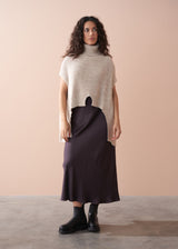 Woman wearing a beige turtleneck sweater and dark purple skirt against a beige background