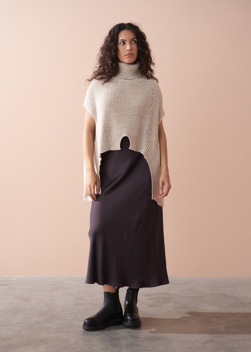 Woman wearing a beige turtleneck sweater and dark purple skirt against a beige background