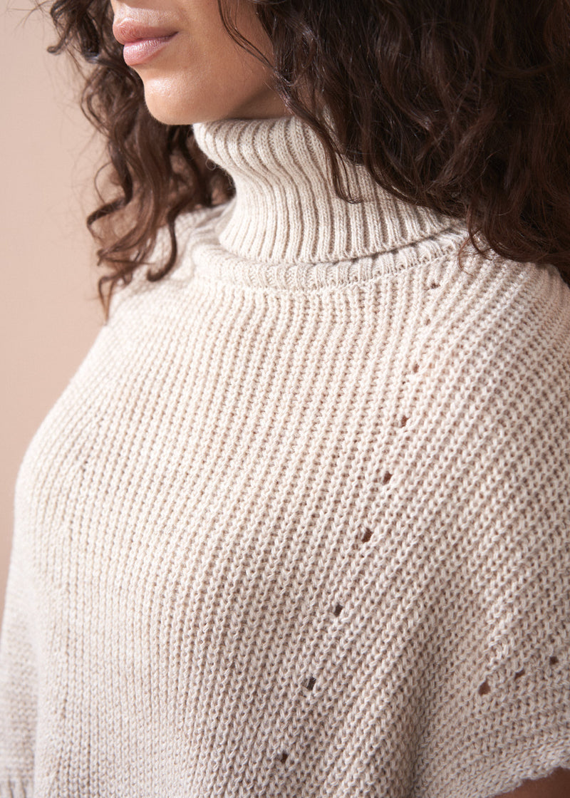 Close-up of a person wearing a beige knitted sweater with a plain background
