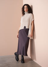 Woman wearing a beige knit poncho over a long dark skirt against a plain background