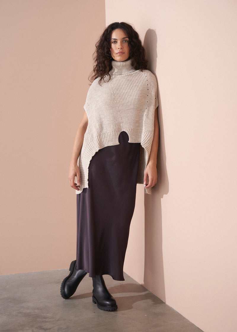 Woman wearing a beige knit poncho over a dark dress against a beige wall.