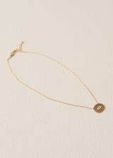 A delicate gold chain necklace with a small circular textured pendant, displayed on a light beige textured background.