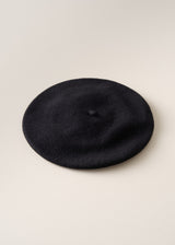 A classic black felt beret hat photographed from a top-down angle on a light beige textured background.
