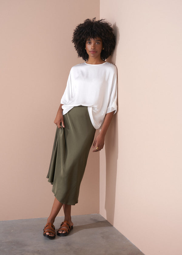Woman wearing a white top and green satin midi skirt against a beige wall.