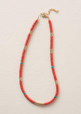 A beaded necklace with coral-red beads, accented with small turquoise beads and gold-toned cylindrical spacers, displayed on a textured off-white background.