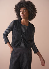 Person wearing a black textured waistcoat over a striped shirt on a beige background