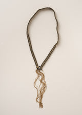 A long beaded necklace with black and beige beads, featuring a central silver-toned medallion and a long, frayed tassel at the bottom, displayed against a plain off-white background.