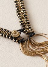 A close-up of a black and gold beaded necklace with a round gold medallion and multiple dangling gold chains on a textured off-white background.