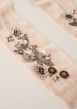 Close-up of a pale blush pink fabric accessory with ornate floral embroidery and metallic embellishments in silver, gold, and dark brown, laid on a light neutral background.