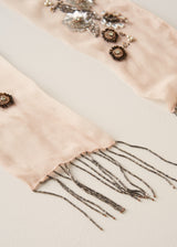 A close-up of a pale blush pink scarf with silver beaded fringe and ornate silver and dark brown floral bead embellishments, laid flat on a light beige background.