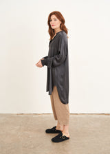 A model wearing a long dark grey satin tunic top with ruched sleeves and a v neck with light brown linen trousers and black clogs