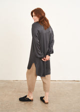 A model wearing a long dark grey satin tunic top with ruched sleeves and a v neck with light brown linen trousers and black clogs