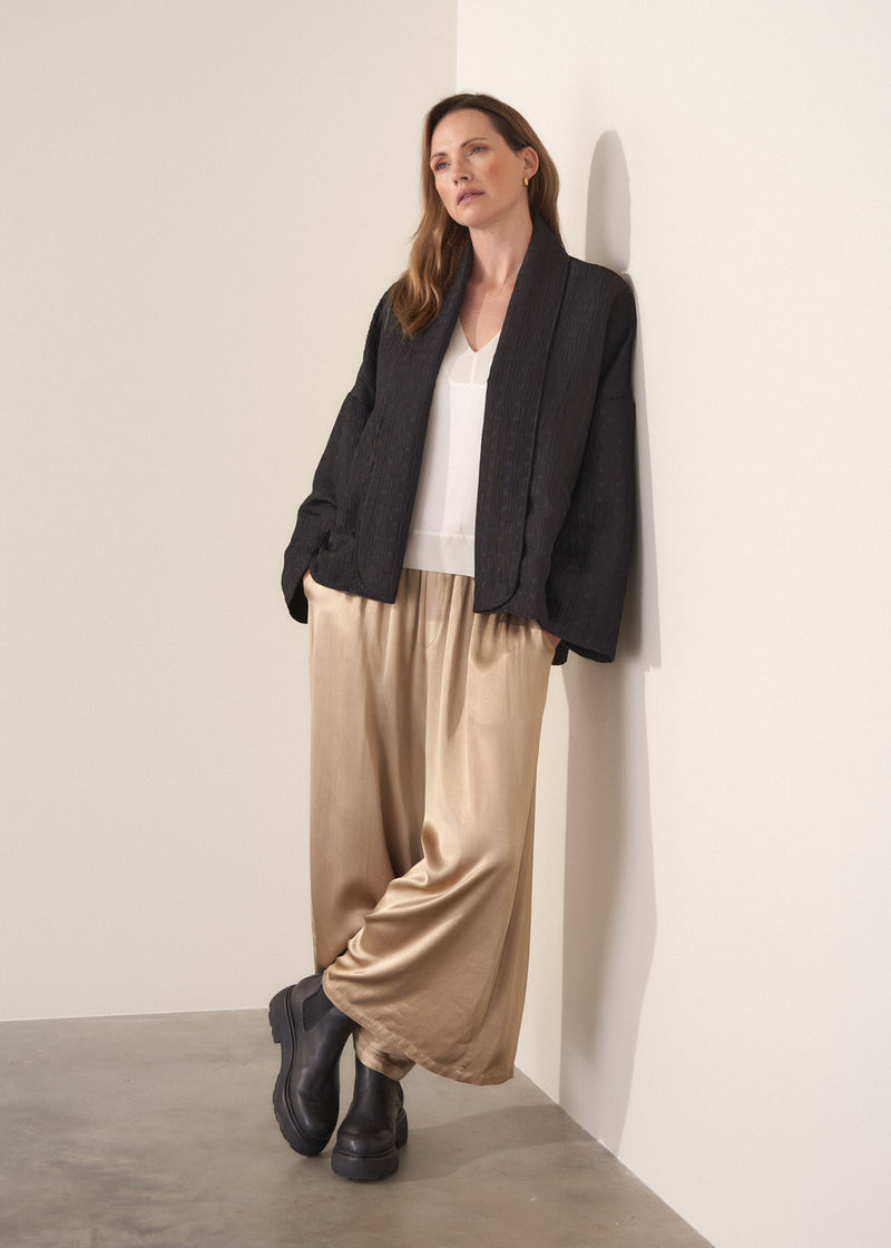 Woman wearing a oversized quilted jacket over a white top and gold trousers against a light background