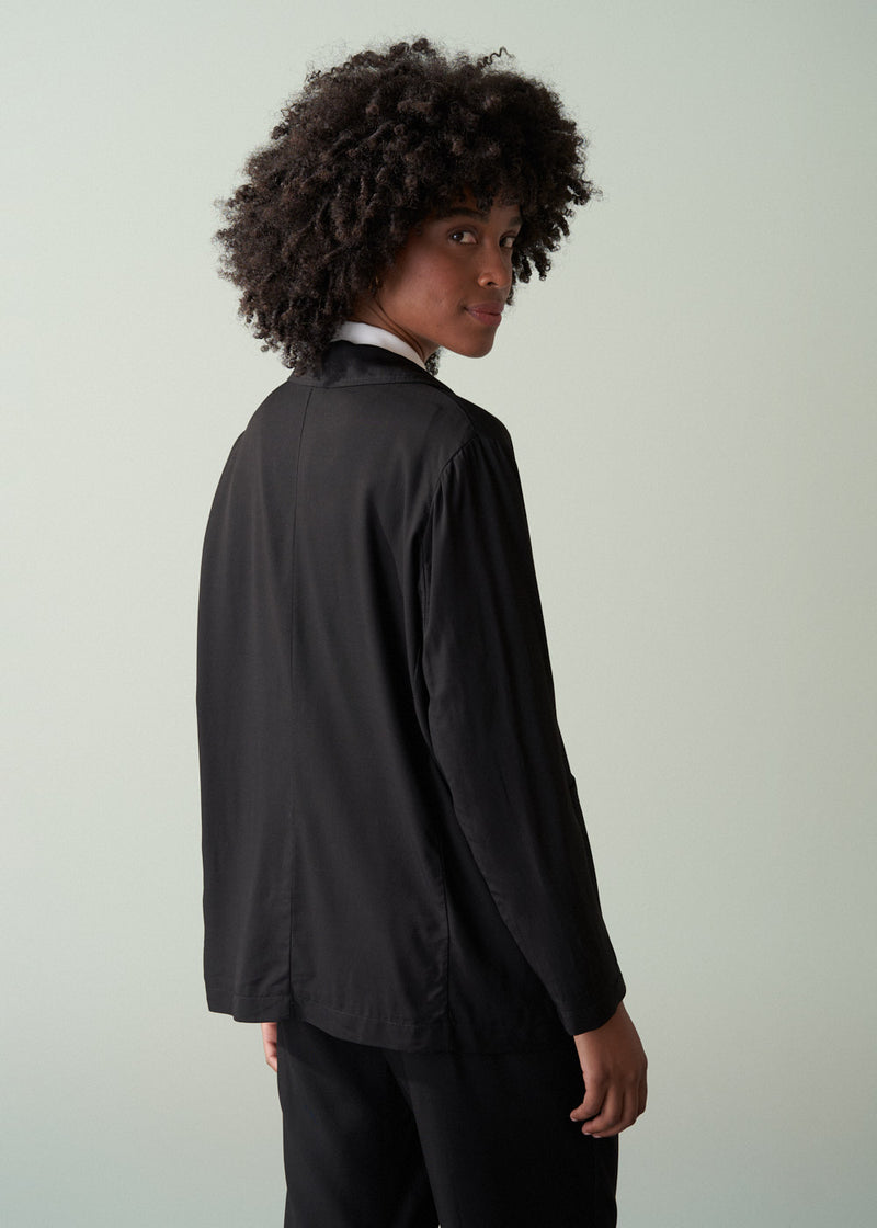 Woman wearing a black relaxed blazer, white shirt and black trousers against a light gray background