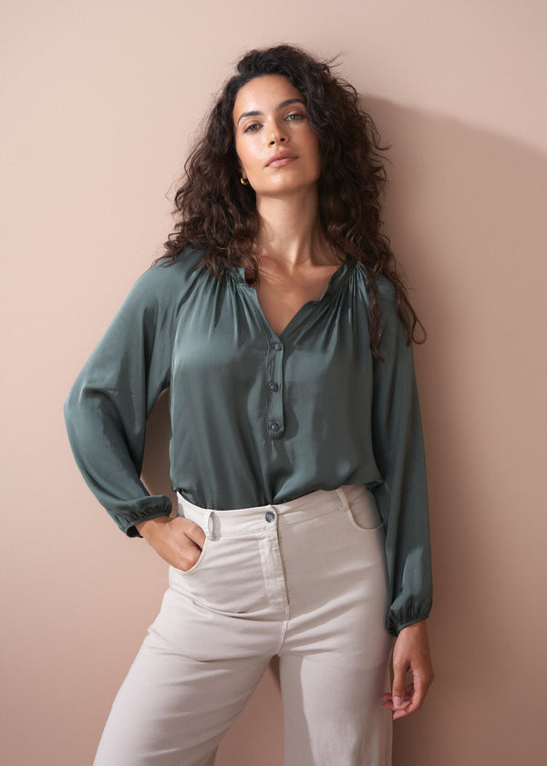Woman wearing a green blouse and off white wide leg trousers against a beige wall