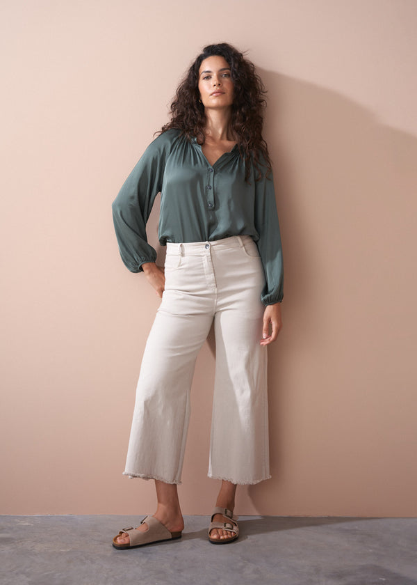 Woman wearing a green blouse and off white wide leg trousers against a beige wall