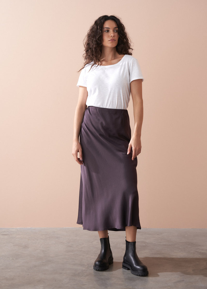 Woman wearing a white t-shirt and dark burgundy satin slip skirt against a beige background
