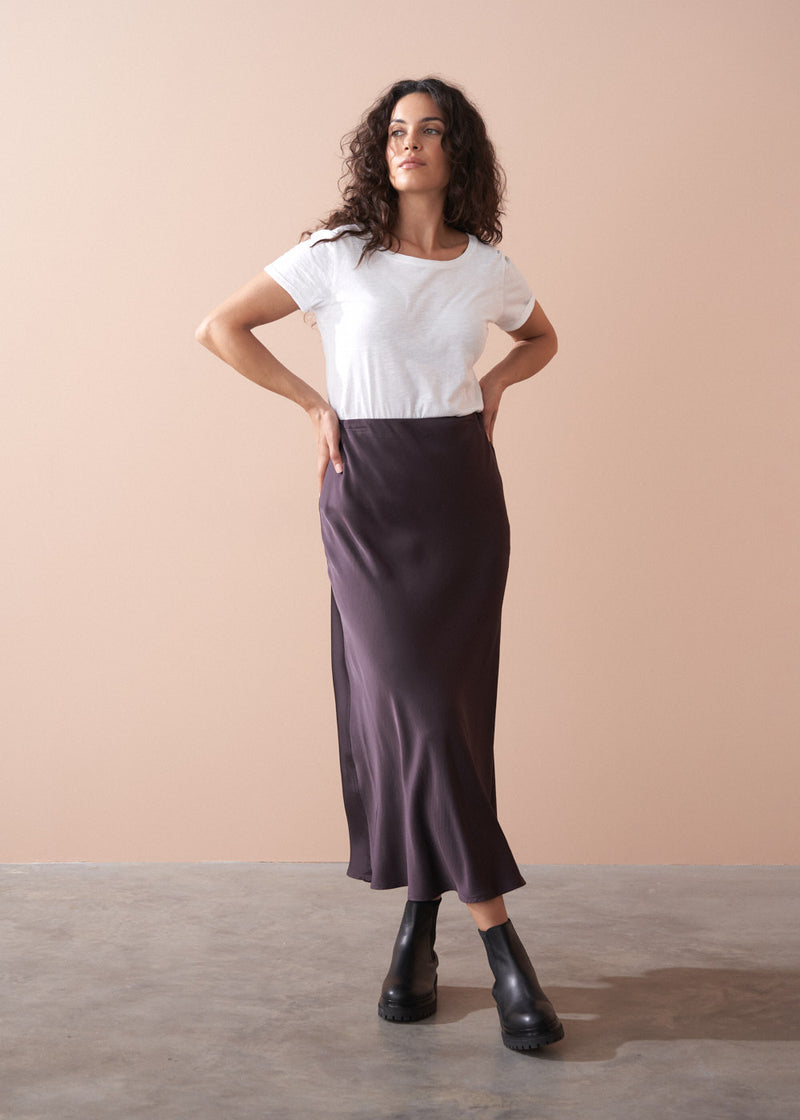 Woman wearing a white t-shirt and dark burgundy satin slip skirt against a beige background