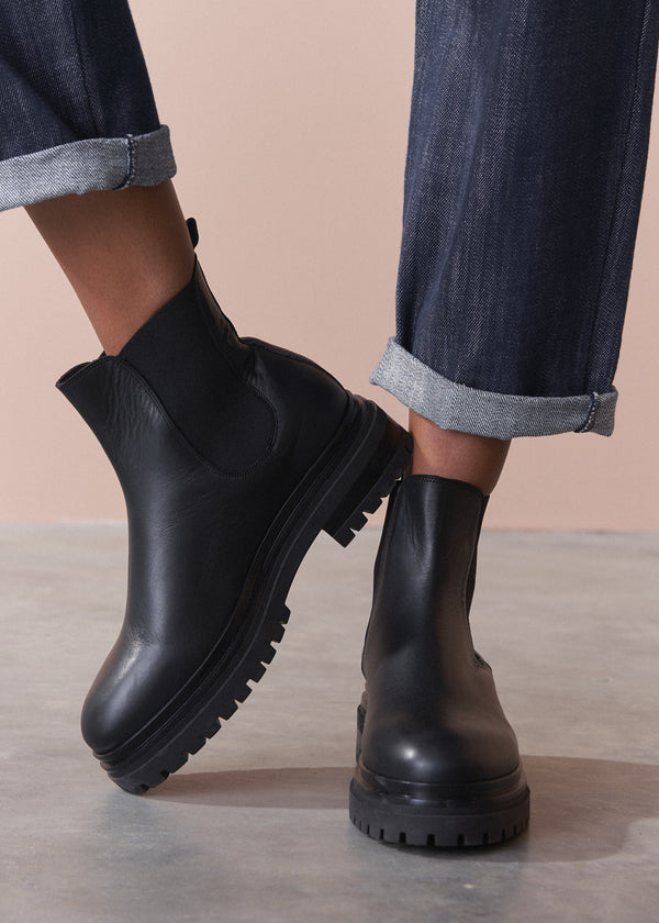 Black leather boots worn with rolled-up jeans on a neutral background