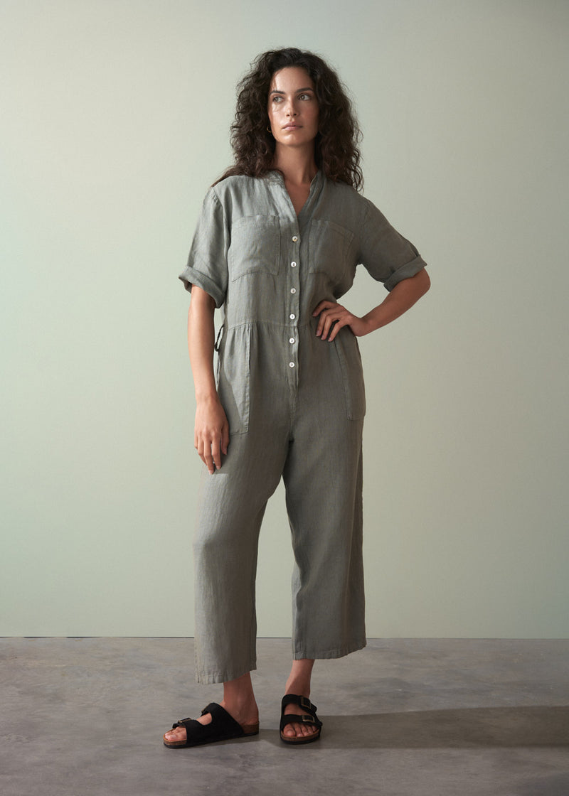 Woman wearing a khaki green linen jumpsuit standing against a plain background