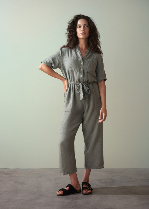Woman wearing a khaki green linen jumpsuit standing against a plain background