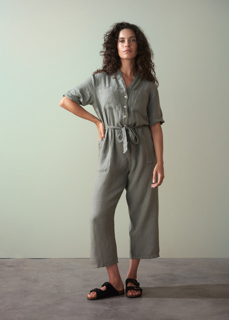 Woman wearing a khaki green linen jumpsuit standing against a plain background