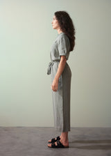 Woman wearing a khaki green linen jumpsuit standing against a plain background
