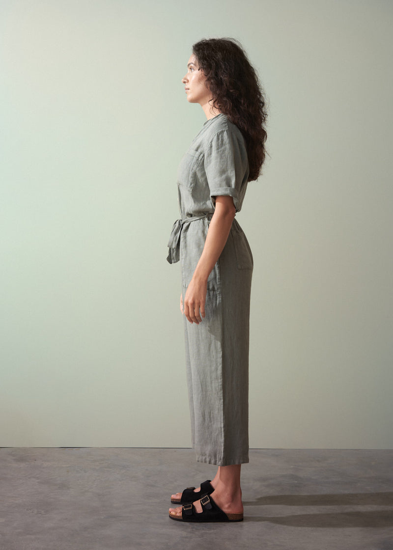 Woman wearing a khaki green linen jumpsuit standing against a plain background