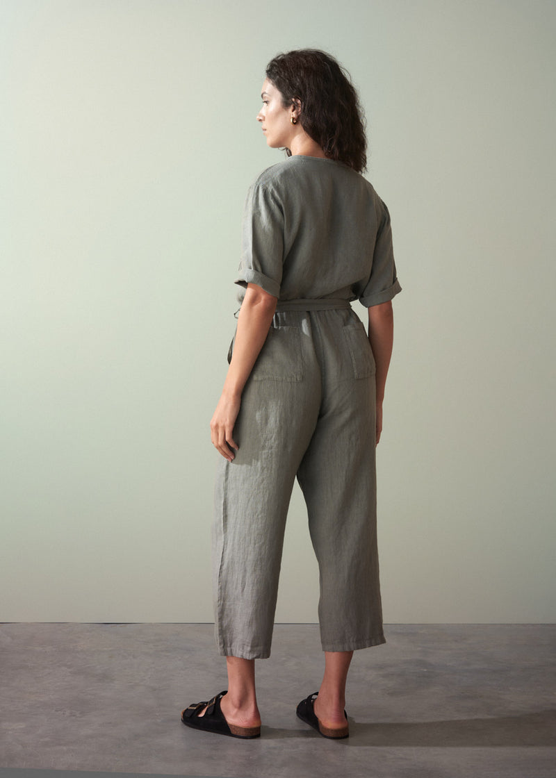Woman wearing a khaki green linen jumpsuit with a belt against a plain background