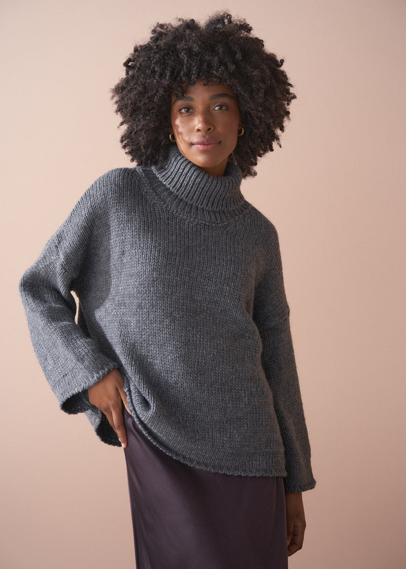 Person wearing a grey chunky knit sweater and satin bias cut skirt against a beige background