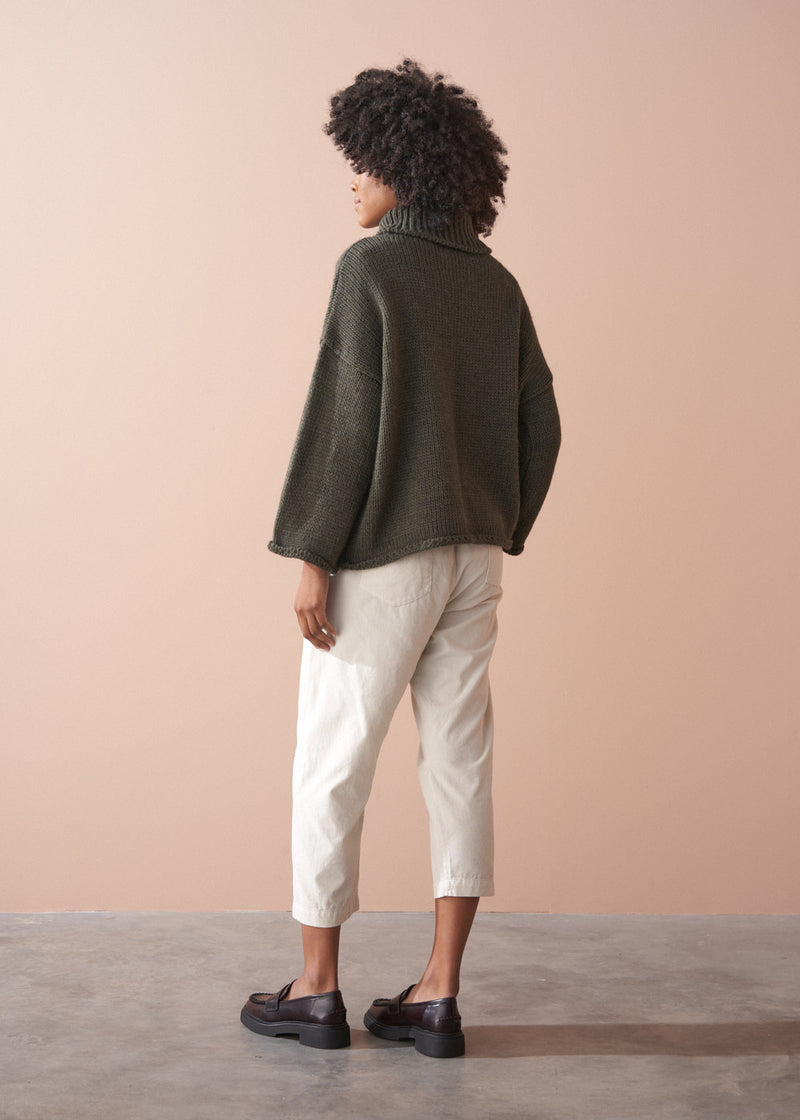 Woman wearing a green chunky knit toll neck sweater and oatmeal cord trousers against a beige background