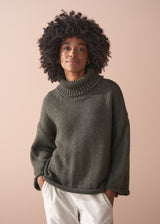Woman wearing a green chunky knit toll neck sweater and oatmeal cord trousers against a beige background
