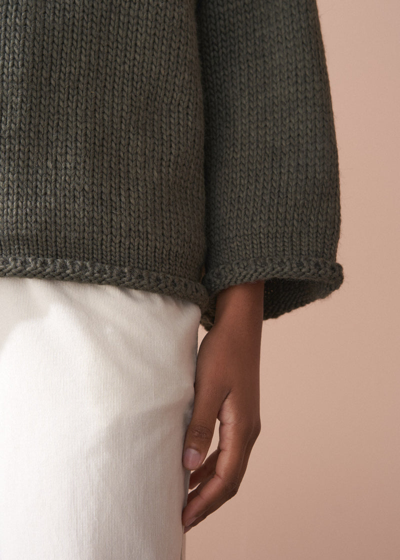 Woman wearing a green chunky knit toll neck sweater and oatmeal cord trousers against a beige background
