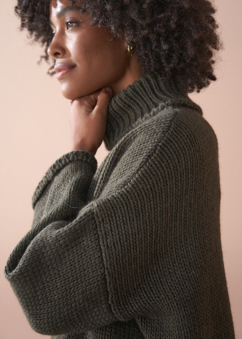 Woman wearing a green chunky knit toll neck sweater and oatmeal cord trousers against a beige background