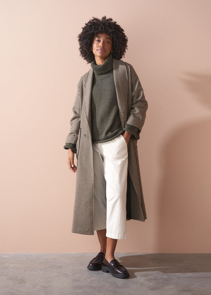 Woman wearing a green chunky knit toll neck sweater and oatmeal cord trousers against a beige background