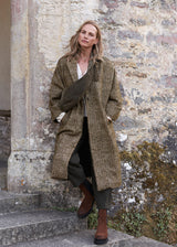 A woman stands on stone steps against a weathered stone wall, wearing a long brown and beige herringbone patterned coat over a white top and dark trousers, with brown ankle boots and a dark shoulder bag.