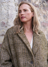 A woman with blonde hair wearing a textured brown and beige plaid coat over a white top, standing against a rustic stone wall.