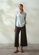 Woman wearing a light blue shirt and brown pants standing against a plain background