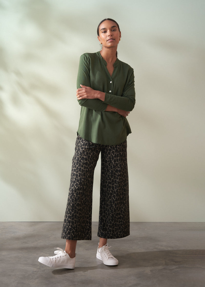 Woman wearing a green shirt and patterned pants standing against a plain background