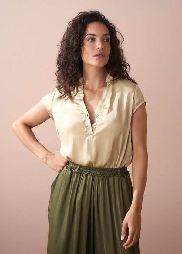 Woman wearing a beige satin blouse and green wide trousers against a plain background