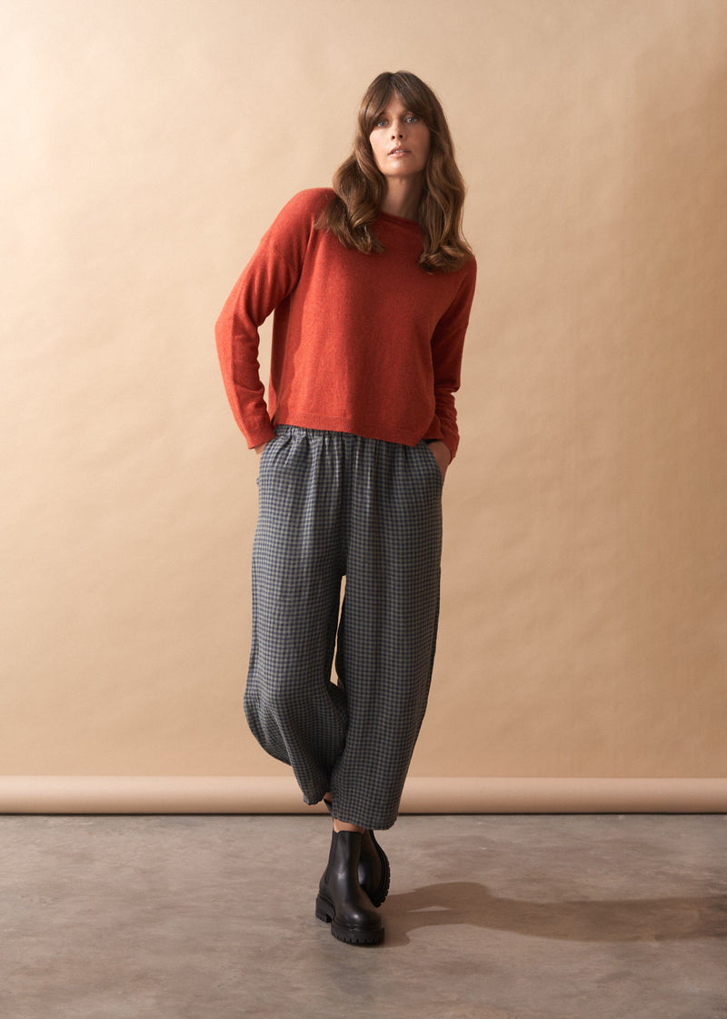 Woman wearing a red sweater and checkered pants against a beige wall.