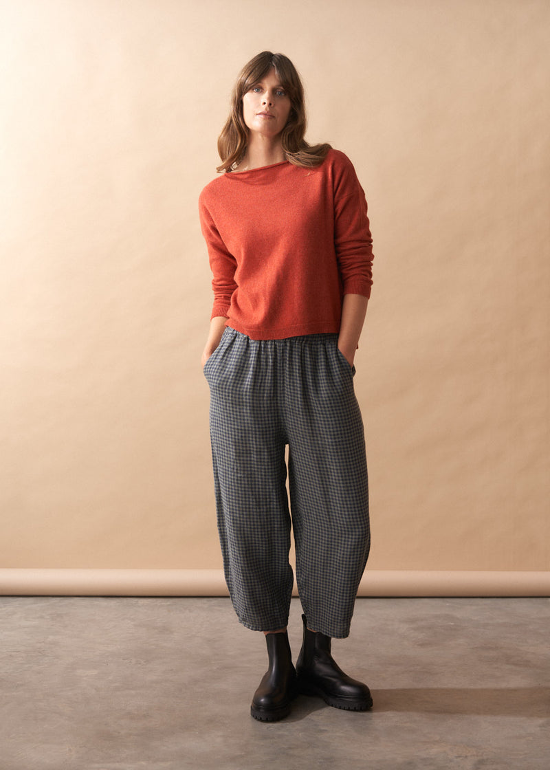Woman wearing a rust-colored sweater and checkered pants against a beige wall.