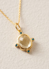 A gold pendant necklace featuring a large, faceted pale yellow stone surrounded by four small round blue gemstones, shown on a textured off-white background.