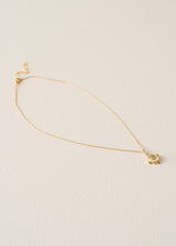 A delicate gold chain necklace with a small, minimalist gold pendant, displayed on a textured off-white background.