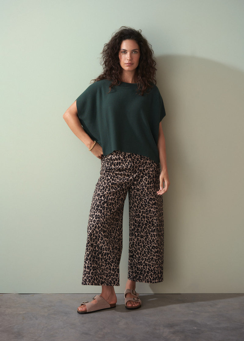 Woman wearing dark green short sleeve knit sweater with leopard print trousers against a plain background.