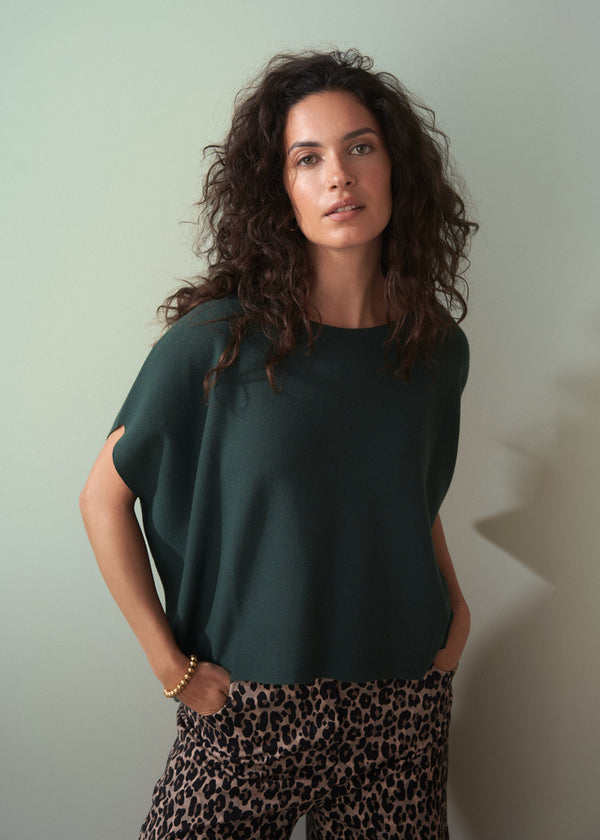 Woman wearing dark green short sleeve knit sweater with leopard print trousers against a plain background.