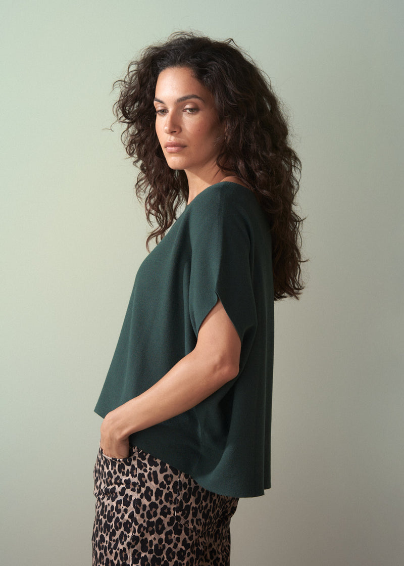 Woman wearing dark green short sleeve knit sweater with leopard print trousers against a plain background.